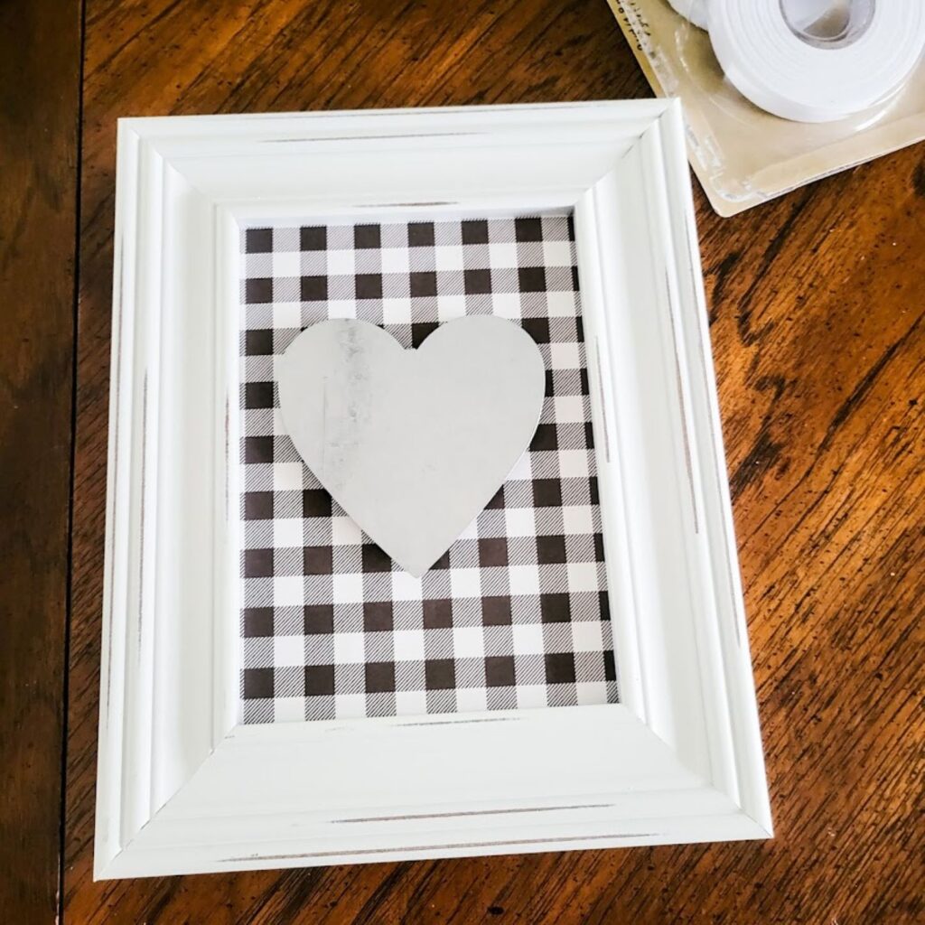 A picture containing a picture frame with a metal heart centered onto the scrapbook paper.