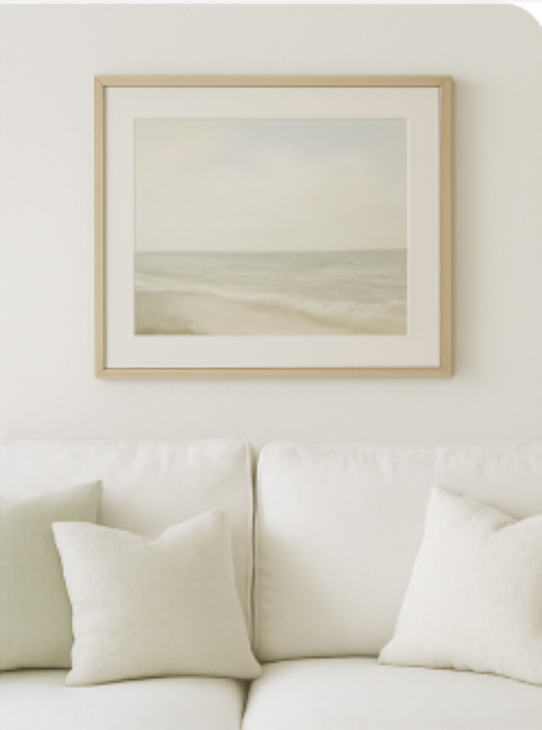 A white living room with a white sofa and throw pillows and a muted seascape painting on the wall how to add a relaxing summertime vibe to your home