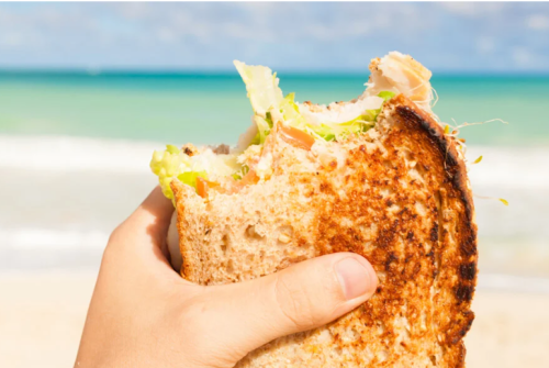 a hand holding a sandwich at the beach your beach grocery list