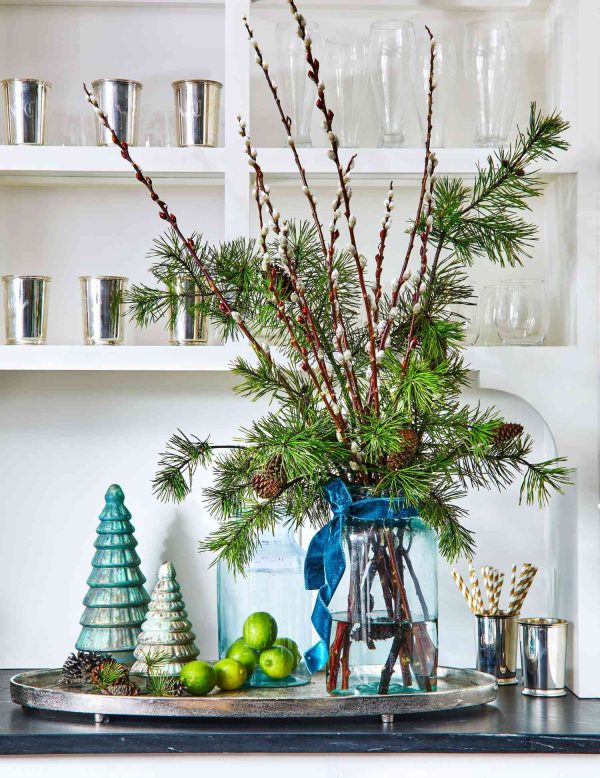 a centerpiece with pine boughs and twigs in a clear glass vase sitting on a table with fruit and mini Christmas trees