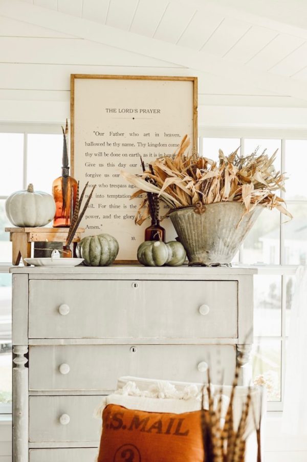 a white dresser with a sign, arrangement of dried flowers and pumpkins on the tabletop